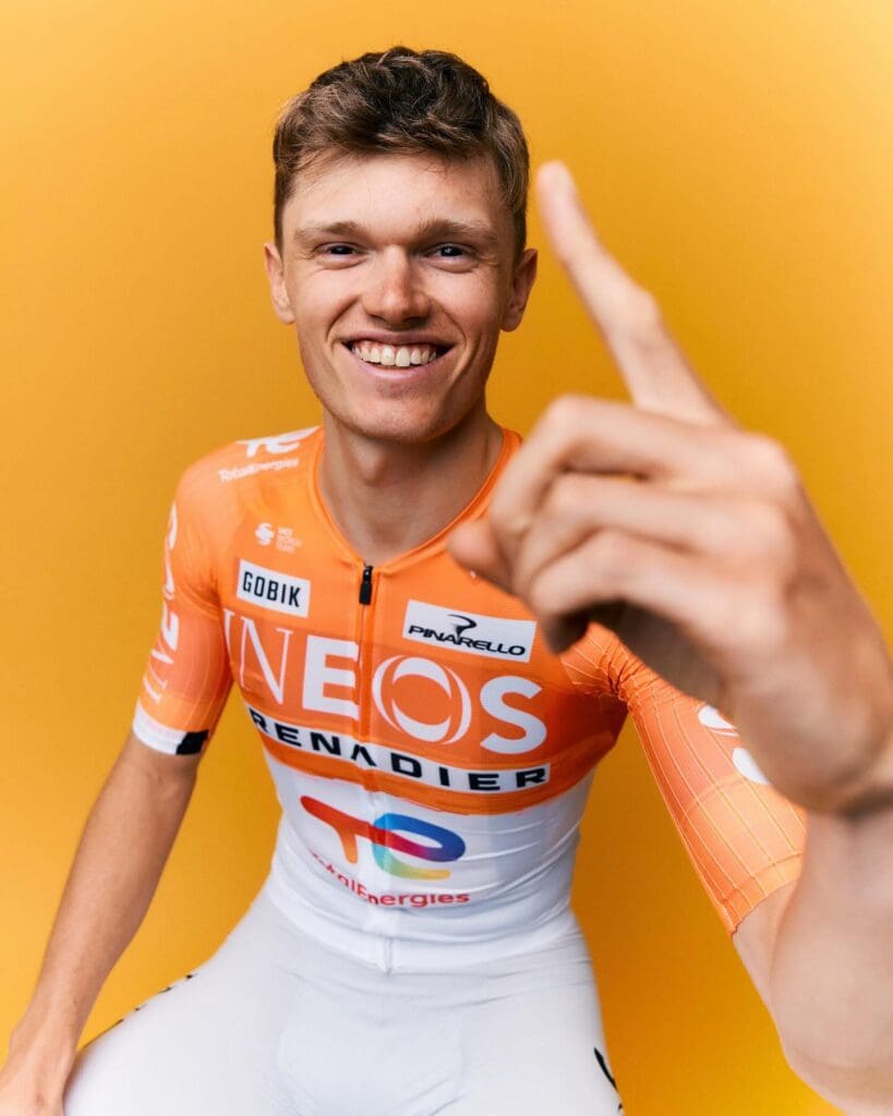 Oscar Onley, short blond-haired cyclist, is pictured in new Ineos kit of orange chest and sleeves and white lycra below. He is smiling and holding up his index finger.